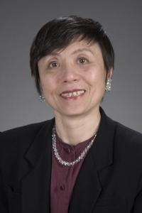 Dr. Ai smiling in a dark purple blouse with black blazer and silver necklace against a grey background