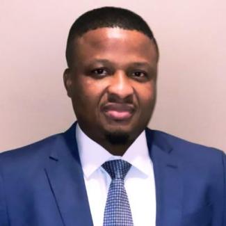 Dr Sorkpor smiling, wearing a blue blazer, white dress shirt, and blue necktie with white grid lines. He appears before a plain beige background.