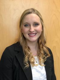 Katharine in front of golden wood paneling, wearing a black suit jacket over a white blouse.  She is smiling and has long blonde hair.