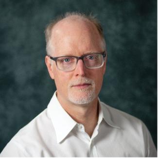 Dr Schmidt is sitting in front of a blue marbled background wearing a white button-up shirt.