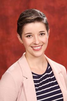Dr. Sheffler smiling wearing a black and white striped shirt and pink blazer against a maroon background