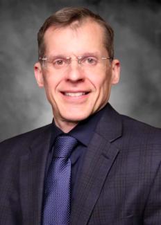 Dr. McGregor smiling wearing glasses a black shirt and blazer with a blue tie against a grey background