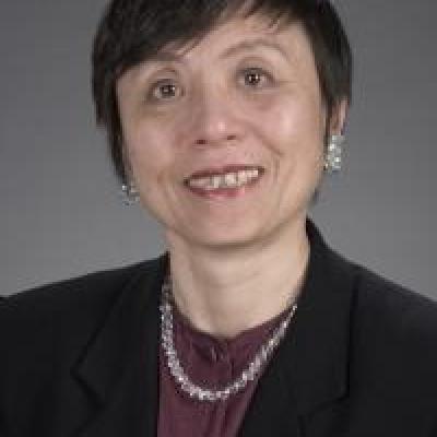 Dr. Ai smiling in a dark purple blouse with black blazer and silver necklace against a grey background