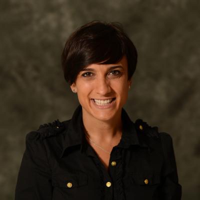 Dr Geraldine Martorella smiling, wearing a black collared blouse with gold buttons. She appears before a neutral toned photo backdrop.