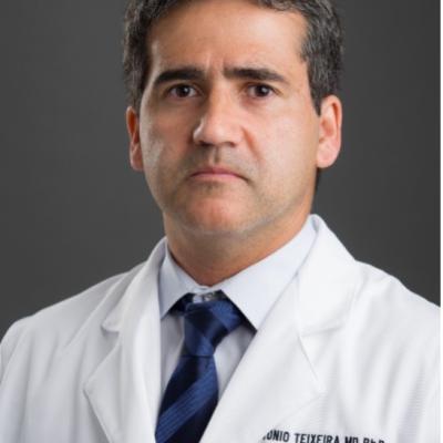 Dr. Teixeira wearing white lab coat and navy tie against dark grey background